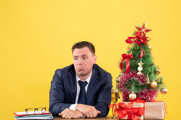 坐着坐在圣诞树旁的桌子旁的一个好奇的男人和黄色的礼物圣诞树桌子办公室