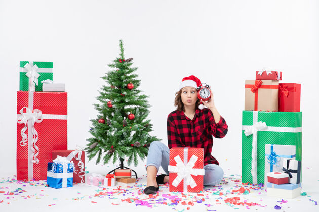 人前视图的年轻女子坐在圣诞礼物周围拿着白色墙上的时钟十二月礼物圣诞树