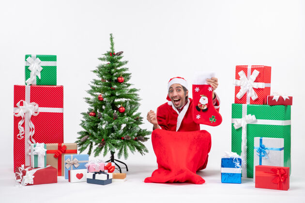 礼物惊喜的圣诞老人正坐在地上 手里拿着圣诞袜 旁边摆放着礼物和装饰在白色背景上的新年树圣诞地面圣诞老人