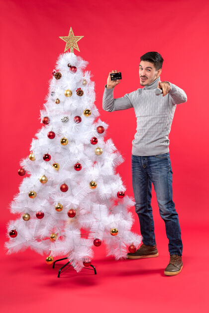 微笑圣诞气氛 微笑的年轻人站在装饰好的圣诞树旁 拿着麦克风和电话 指着前方站着的人装饰小伙子抱着