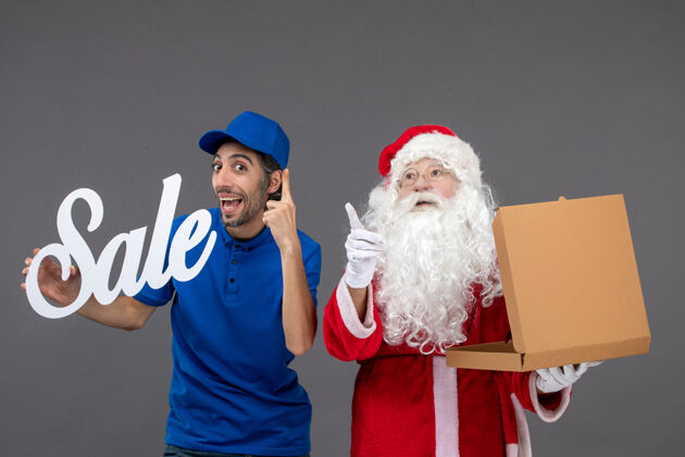 快递圣诞老人的正面图 男性快递员拿着销售字条 灰色墙上放着食品盒圣诞老人肖像吉祥物