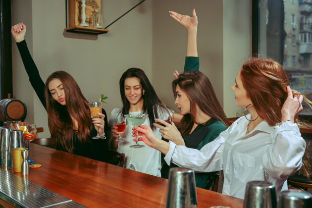 碰杯女朋友们在酒吧喝酒 她们坐在一张摆着鸡尾酒的木桌旁 她们在碰杯胜利饮料鸡尾酒