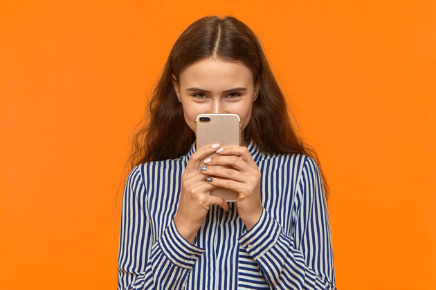智能手机穿着条纹衬衫的快乐积极的年轻女性拿着手机盯着自己的脸 盯着相机短信新闻电话