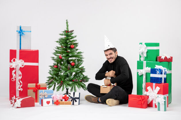 十二月年轻人坐在白色墙上的节日礼物周围的正面视图情感礼物树