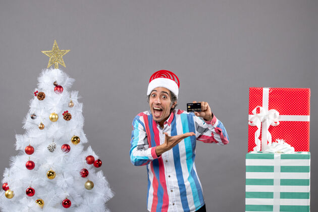 显示前视图快乐的年轻人在圣诞树旁展示他的信用卡灰色信用卡卡