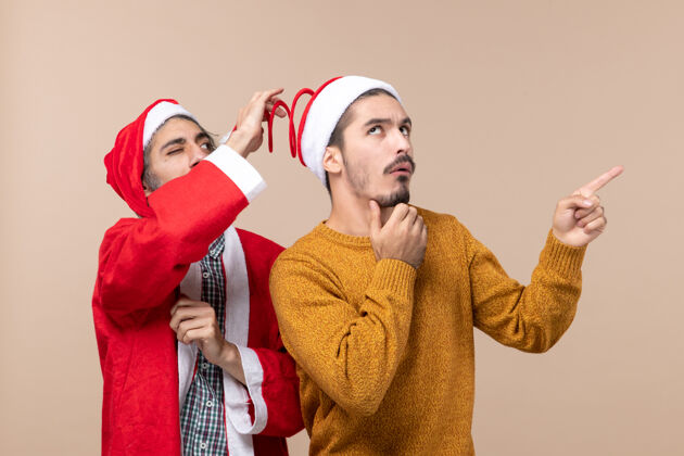 前面正面图两个戴着圣诞帽的朋友 一个试图拿着帽子 另一个在米色孤立的背景上显示方向视图微笑帽子