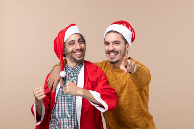 成人前视图两个戴着圣诞帽的朋友在米色孤立的背景下看着相机前面表情米色