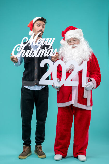 2021年前视图圣诞老人与年轻的男性举行圣诞快乐和蓝色背景上的作品圣诞老人男性举行