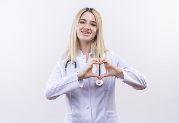 微笑微笑的医生 年轻的金发女孩 戴着听诊器 穿着医用长袍 戴着牙套 在孤立的白色背景上展示着心脏的姿态心脏医学长袍