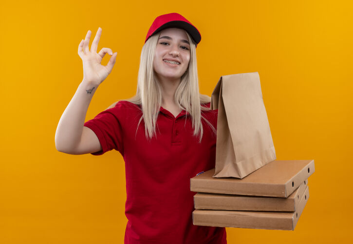 好的微笑的送货年轻女孩穿着红色t恤 戴着帽子 戴着牙套 手里拿着披萨盒和纸口袋 在孤立的橙色背景上显示出okey手势盒子手势口袋