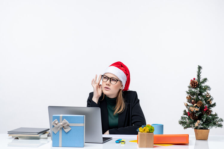 迷人的女人办公室里 一个戴着圣诞老人帽子的漂亮女人坐在桌子旁 桌上放着圣诞树和礼物 心情很混乱 新年快乐女人坐着桌子