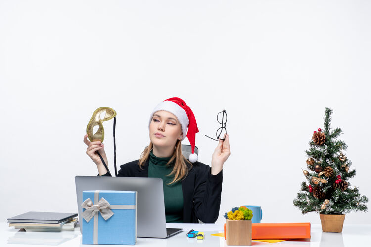 面具在白色背景的办公室里 一位自信的年轻女士戴着圣诞老人帽 眼镜和面具 坐在一张桌子旁 桌子上放着圣诞树和礼物女商人年轻女人礼物