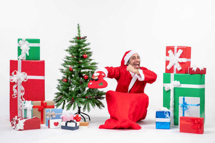 地面新年气氛与有趣的圣诞老人坐在地上 穿圣诞袜子附近的礼物和装饰圣诞树上的白色背景坐着礼物袜子