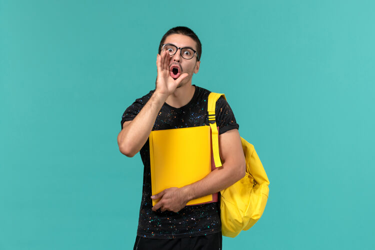 持有身穿深色t恤黄色背包的男学生正面图 手持不同的文件 在浅蓝色的墙上呼喊大学漂亮模特