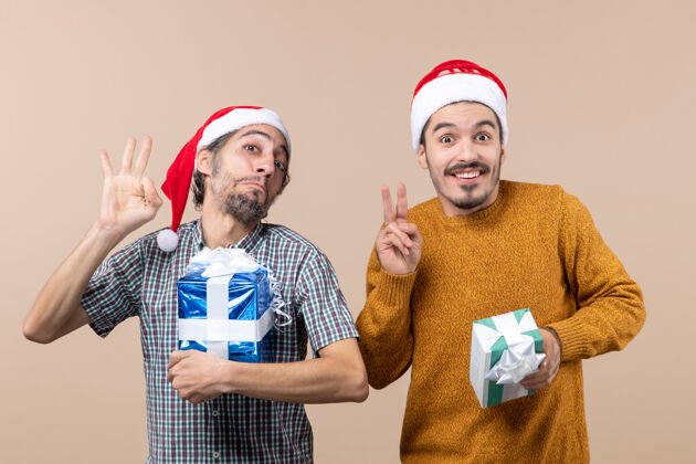 微笑正面图两个幸福的家伙一个做奥基标志 另一个胜利标志 并举行在米色孤立的背景圣诞礼物成人手持两个