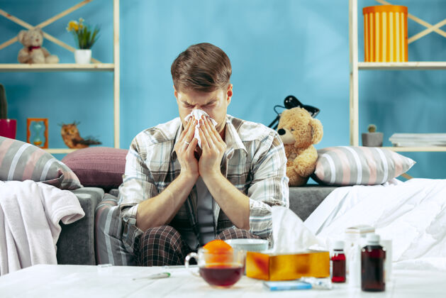 年轻人留着胡子的病人坐在家里的沙发上擤鼻涕冬天 疾病 流感 疼痛的概念在家放松医疗保健的概念体温计发烧流感