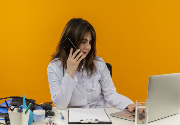 思考想一想中年女医生穿着医用长袍带听诊器坐在办公桌上用笔记本电脑带着医疗工具讲电话 用笔记本电脑在孤立的橙色背景上有复印空间女性工作电话