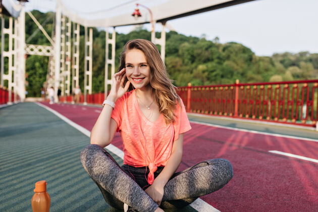 夏天穿着粉色t恤的浪漫年轻女子在训练前摆姿势灵感四射的女模特双腿交叉坐在煤渣跑道上的户外照片苗条跑步者排毒