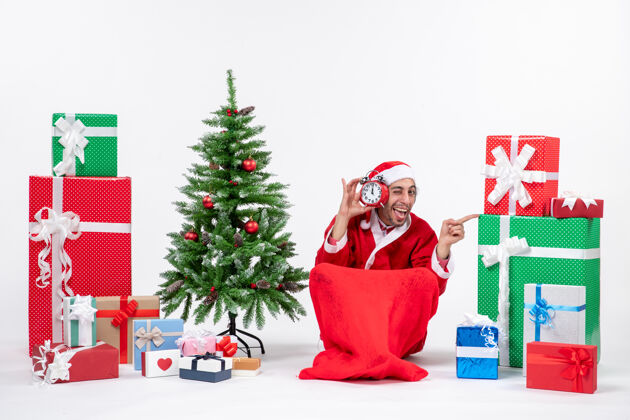 圣诞老人喜怒无常的圣诞老人坐在地上 在礼物和白色背景上装饰圣诞树旁展示钟表圣诞老人装饰地面