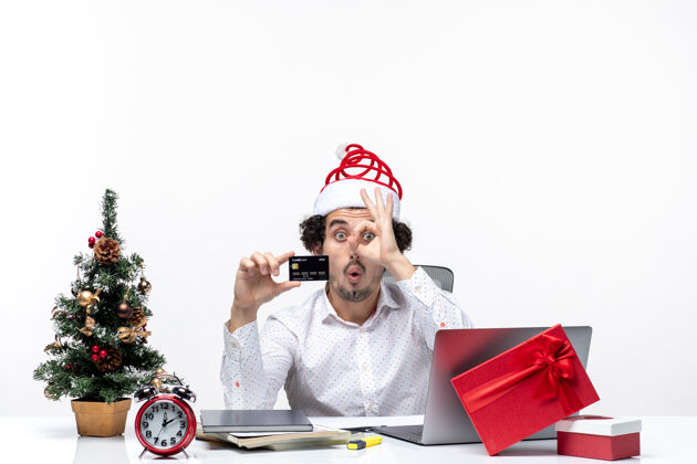 笔记本电脑在白色背景的办公室里 戴着圣诞老人的帽子 拿着银行卡的年轻有趣的商务人士带着节日的喜庆气氛办公室帽子圣诞老人
