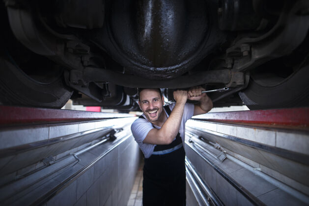 工具微笑的汽车修理工手持扳手在汽车修理厂的卡车下工作的画像人员测试设备