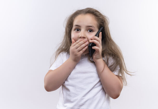女孩惊讶的小女生穿着白色t恤 在孤立的白色背景下用手机捂着嘴说话说话穿着嘴巴