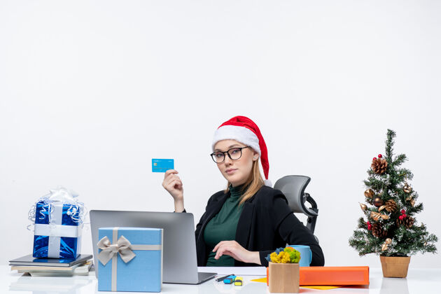圣诞节自信迷人的女人戴着圣诞老人的帽子 戴着眼镜坐在桌子旁 手里拿着银行卡在办公室里电脑公司办公室