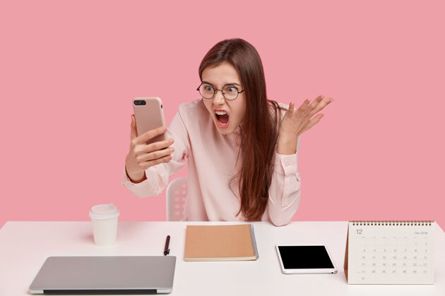自由职业者愤怒恼怒的女人手持现代手机 打视频电话 和同事争吵 在工作场所摆姿势平板电脑短信饮料
