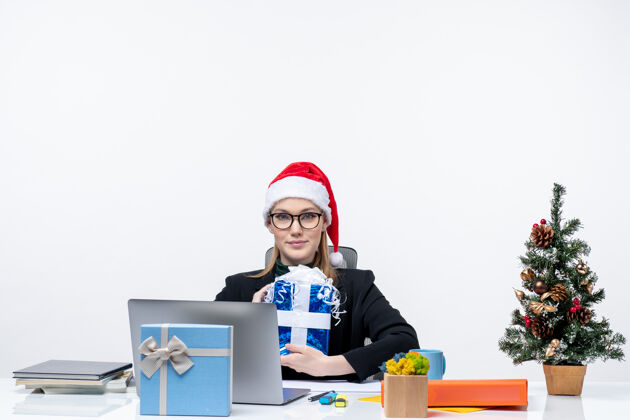 抱着圣诞气氛与戴着圣诞老人帽子和眼镜的年轻女子坐在一张桌子旁 拿着她的礼物自豪地在白色背景眼镜坐着年轻女人