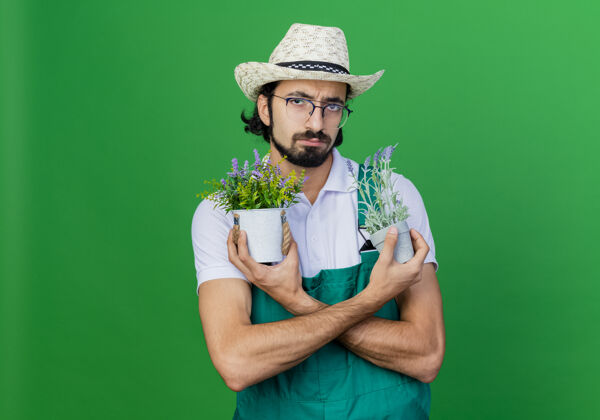 盆栽年轻的留着胡子的园丁 穿着连体衣 戴着帽子 手里拿着盆栽植物 满脸愁容园丁帽子胡子