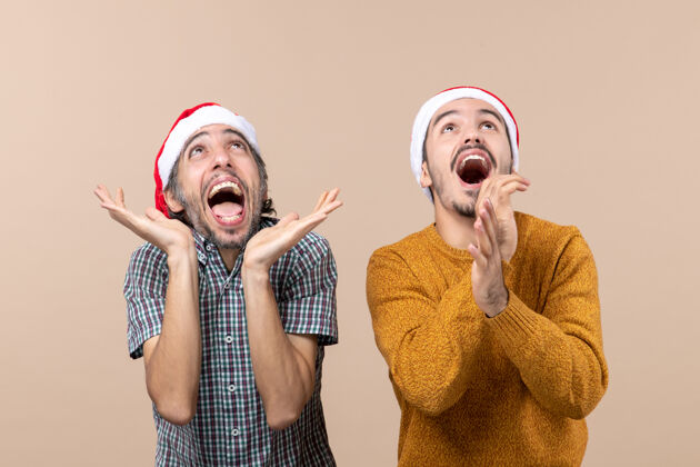脸正面图两个戴着圣诞帽的兴奋的家伙站在米色孤立的背景上拍手面具表情前面