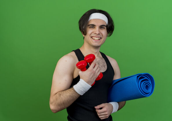 男士身穿运动服 头戴头巾 手持哑铃和瑜伽垫的年轻运动型男子笑容可掬哑铃运动装站着