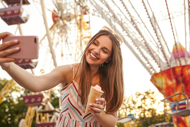 娱乐年轻漂亮的长发黑发女士穿着浪漫的夏装 一边用手机自拍一边在景点上空摆姿势 手里拿着冰激凌筒 开心地微笑着温暖情绪女人