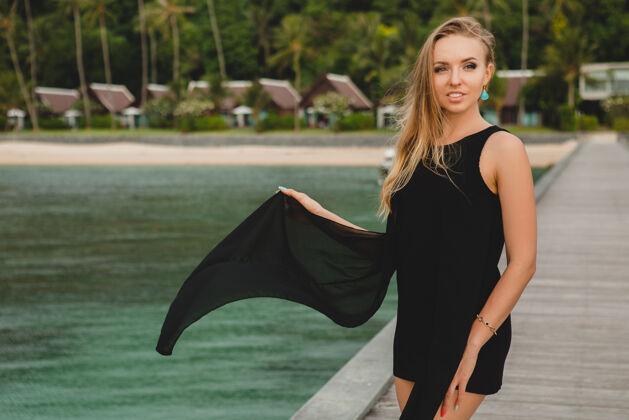 风穿着黑色连衣裙的美丽迷人的女人在豪华度假酒店的码头上摆姿势 暑假 热带海滩幸福度假胜地热带