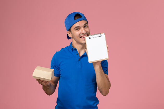 男人正面图身穿蓝色制服披肩的年轻男性快递员手持小快递食品包和笔记本在浅粉色的墙上送货包裹送货