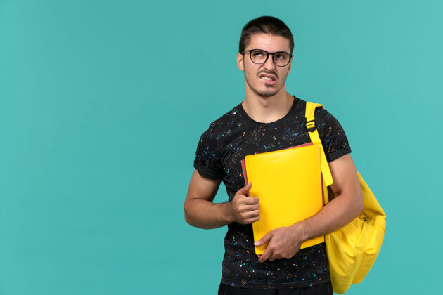 大学身穿深色t恤黄色背包的男学生正面图 浅蓝色的墙上放着不同的文件思考成人正面思维