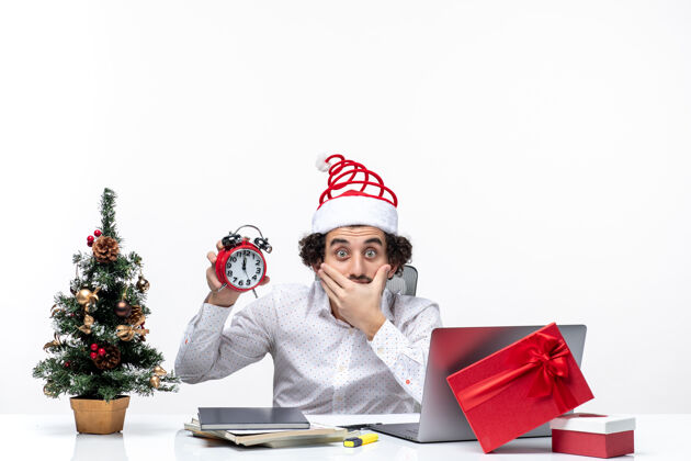 年轻戴着圣诞老人帽的年轻商务人士 带着闹钟 坐在白色背景的办公室里仔细思考着一些事情东西商务人士商务