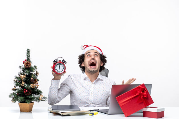 坐着年轻愤怒的商人戴着圣诞老人的帽子 拿着钟 坐在黑暗的背景下的办公室里圣诞老人帽子男