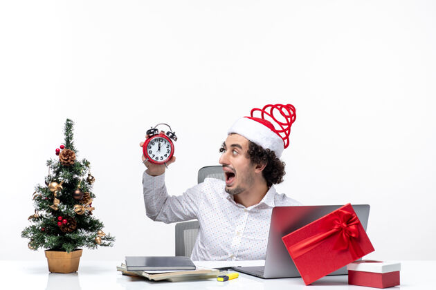 商务人士年轻有趣 情绪激动的商务人士戴着圣诞老人的帽子 拿着一个白色背景的时钟坐在办公室里男性情绪化圣诞节