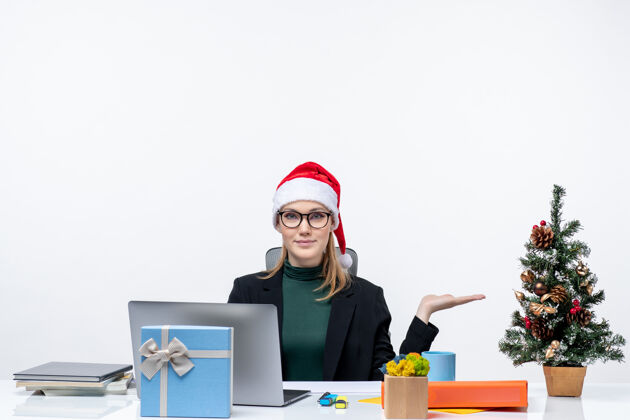 礼物一位微笑的商务女士 戴着圣诞老人帽 坐在一张桌子旁 桌上放着圣诞树和礼物 指着办公室左侧白色背景上的东西一边指着帽子