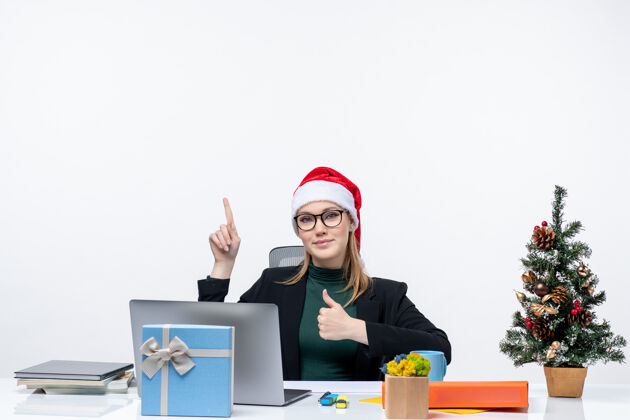 好的一位自信的商务女士 戴着圣诞老人帽 坐在一张桌子旁 桌上放着圣诞树和一份礼物 做着“ok”的手势 在白色背景上指着办公室里的东西圣诞老人礼物桌子