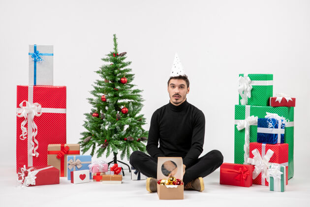 寒冷年轻人坐在白色墙上的节日礼物周围的正面视图礼物人前面