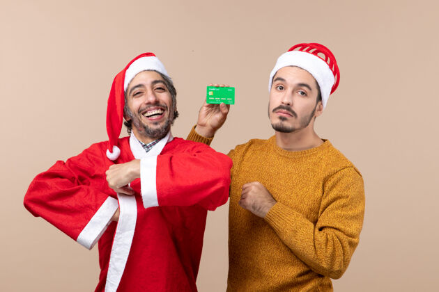 卡片前视图两个家伙一个穿着圣诞老人的外套微笑着 另一个拿着信用卡看着米色背景上的相机两个微笑男人