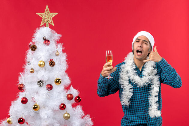 男人圣诞节心情与情绪激动的年轻人圣诞老人帽子在一个蓝色条纹衬衫拿着一杯葡萄酒 并作出了圣诞树附近打电话给我的手势圣诞老人家伙圣诞