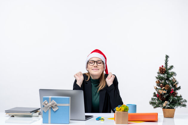 帽子新年的心情与梦想迷人的女人坐在一个圣诞老人的帽子与圣诞树和一个礼物 它在办公室的桌子上女商人圣诞老人圣诞老人