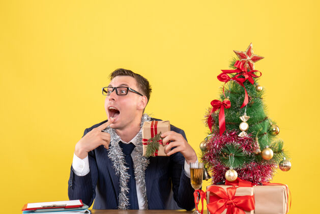 歌手年轻人张着嘴拿着礼物坐在圣诞树旁的桌子上 黄色的礼物礼物礼物桌子