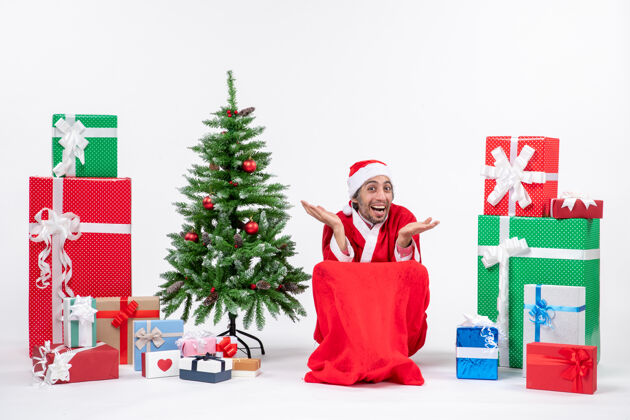 微笑好奇 快乐 情绪化的年轻人打扮成圣诞老人 拿着礼物 在白色背景上装饰圣诞树年轻人年轻人人