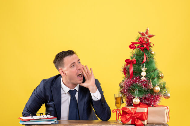 某人年轻人打电话给坐在圣诞树旁桌上的人 并在黄色的礼物上办公室黄色坐着