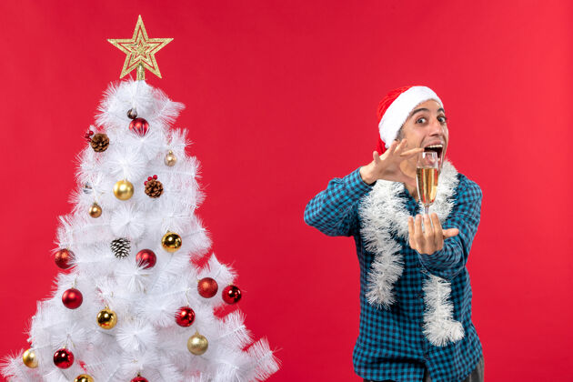 帽子圣诞心情快乐的年轻人戴着圣诞老人的帽子 穿着蓝色的衬衫 在圣诞树旁举起一杯葡萄酒红色年轻人香槟