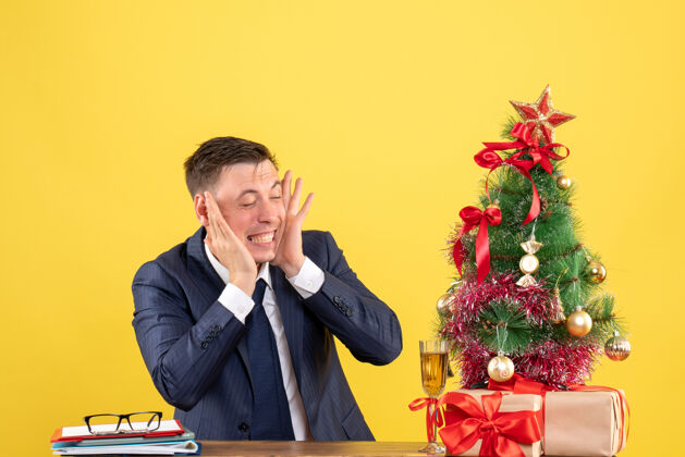 西装年轻人用手指合上耳朵坐在圣诞树旁的桌子旁 用黄色的礼物送人礼物男人年轻人